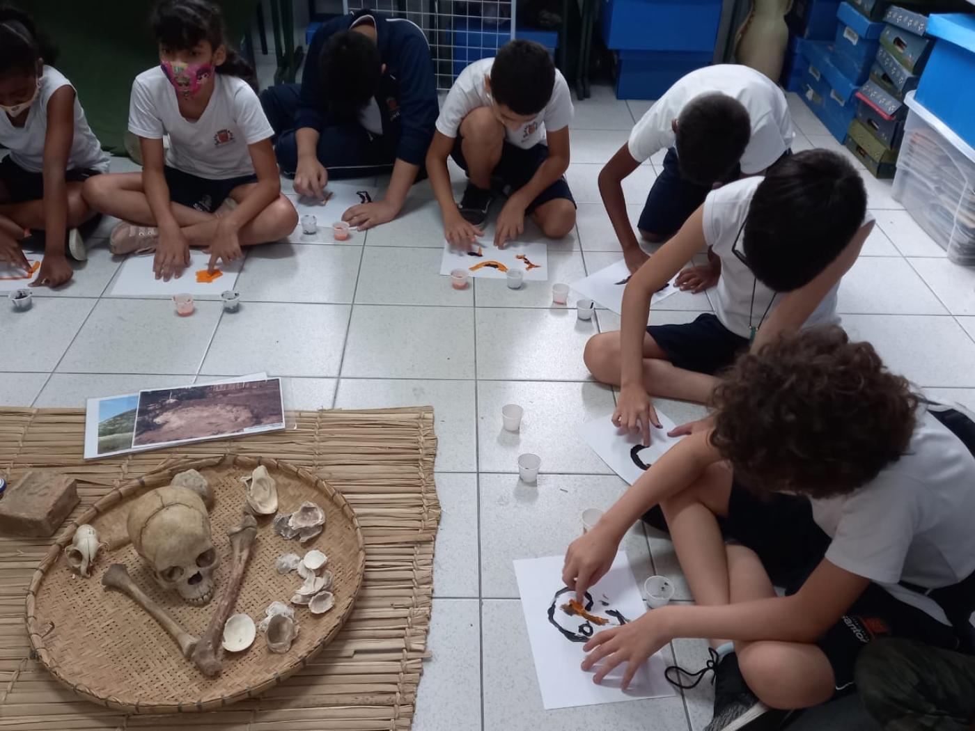 alunos estão sentados no chão desenhando. todos estão diante de cestos com crânios, ossos e outros objetos. #paratodosverem