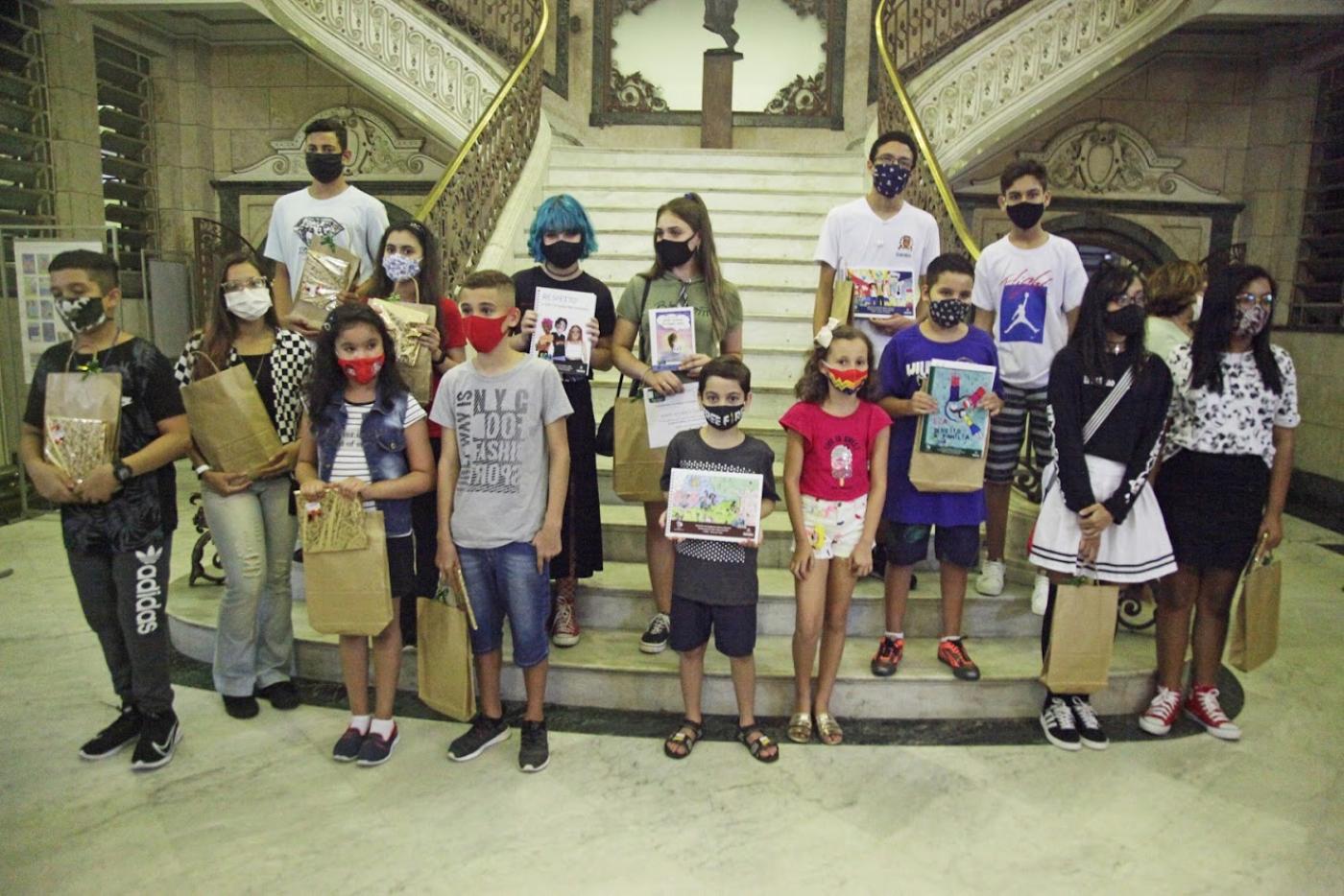 Vencedores de concurso posam para foto no saguão da prefeitura segurando os cadernos com as respectivas capas. #Paratodosverem