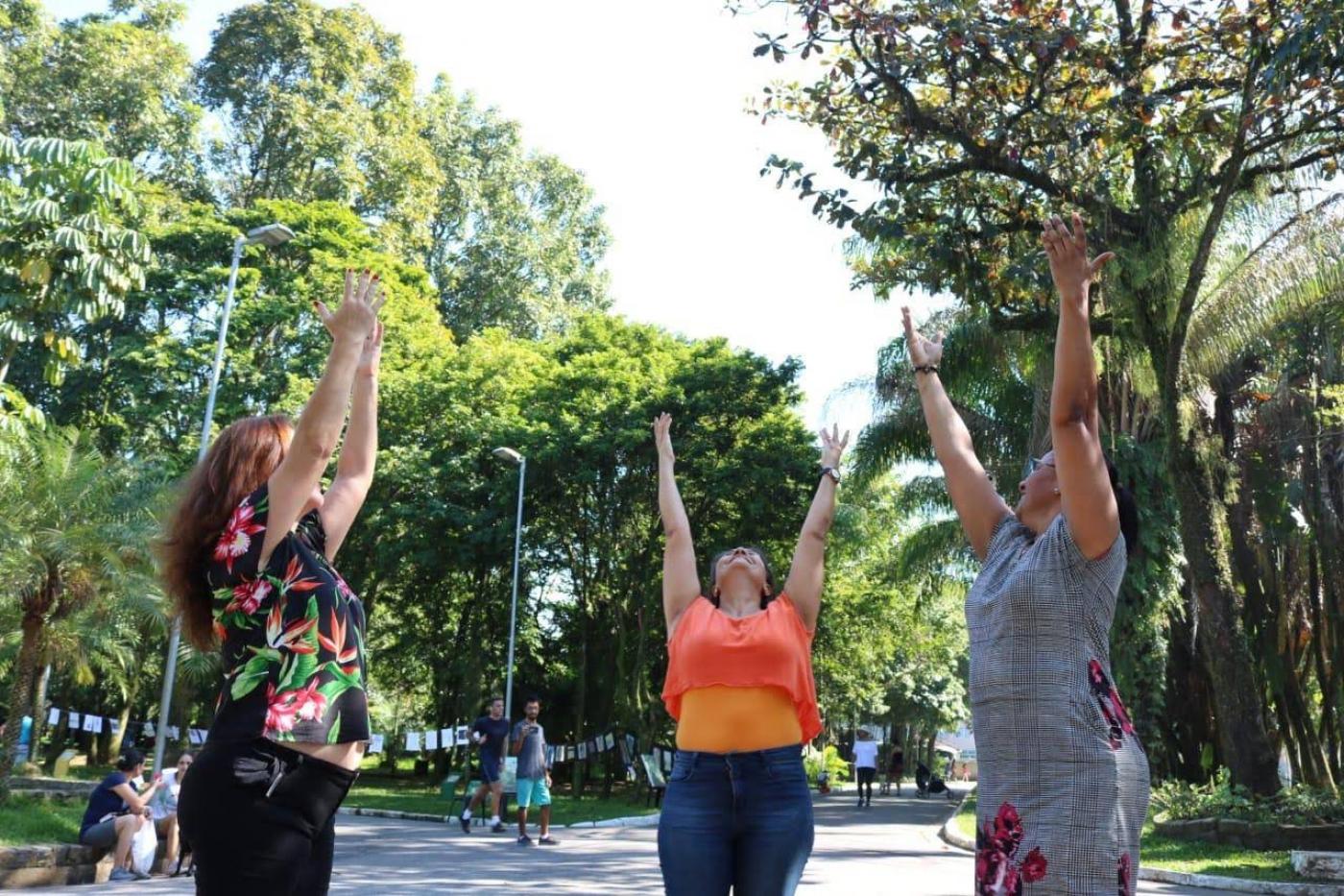 Três mulheres estão em pé em meio a vegetação de parque. Elas estão com os braços para cima, como num movimento de ioga, de inspiração. Olham para o alto. #Pracegover