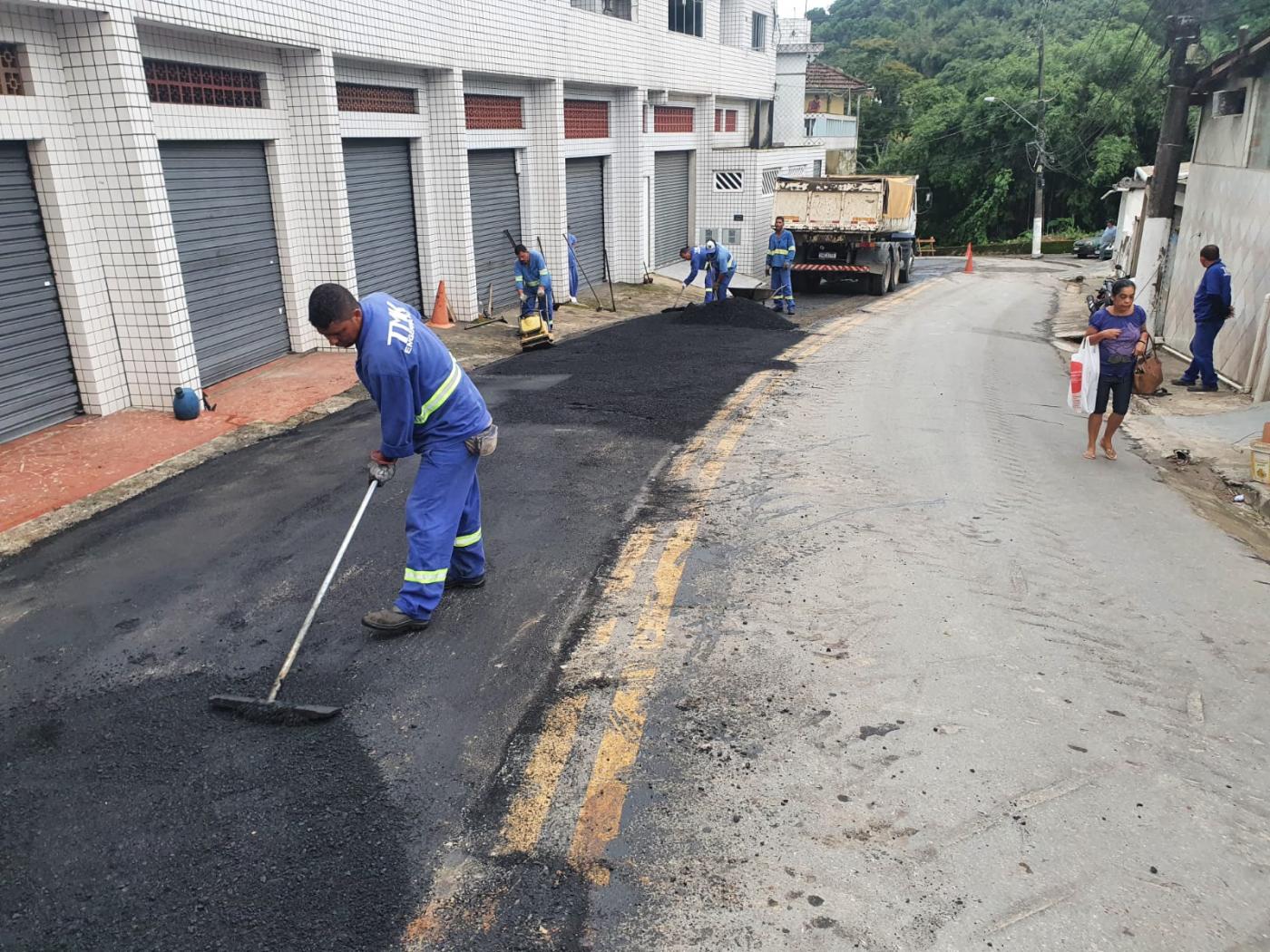 trecho de rua é asfaltado. Há homens ajustando o asfalto e a máquina compactadora ao fundo. #paratodosverem