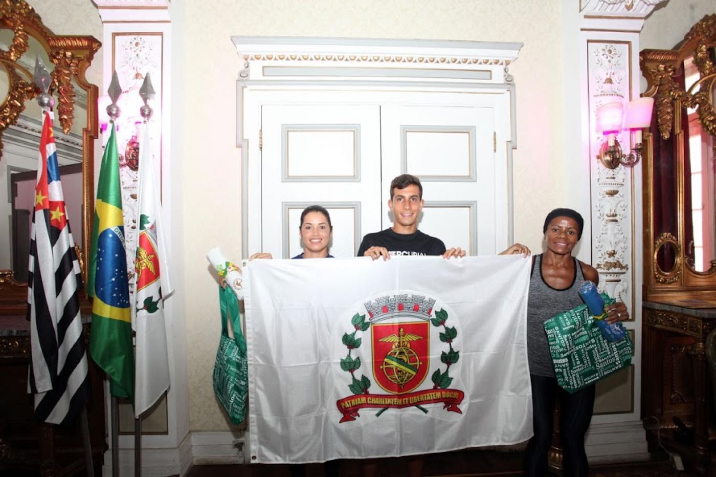 Os três atletas seguram bandeira da Cidade de Santos no salão nobre da prefeitura. #pracegover