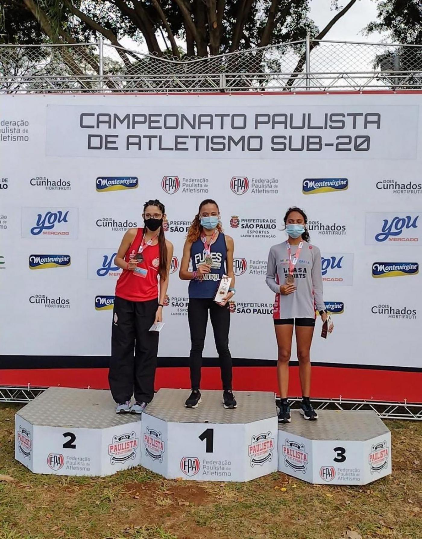 Caroline de Oliveira Gomes está no primeiro lugar do pódio, ao centro, entre outras duas atletas. #paratodosverem