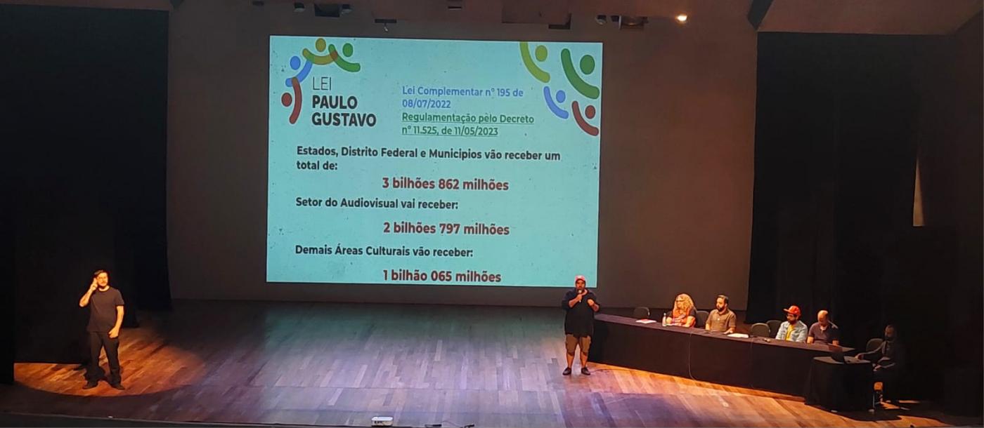 duas pessoas em pé em um palco. Uma projeção ao fundo com texto e quatro pessoas sentadas a uma mesa também no palco. #paratodosverem 