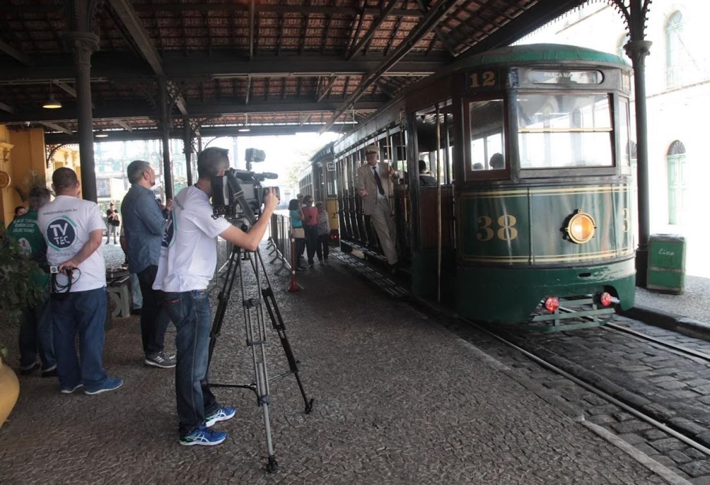 Equipe está na estação de bonde pegando imagens do bonde que está parado. Um dos homens manuseia a câmera que está montada em tripé. #paratodosverem