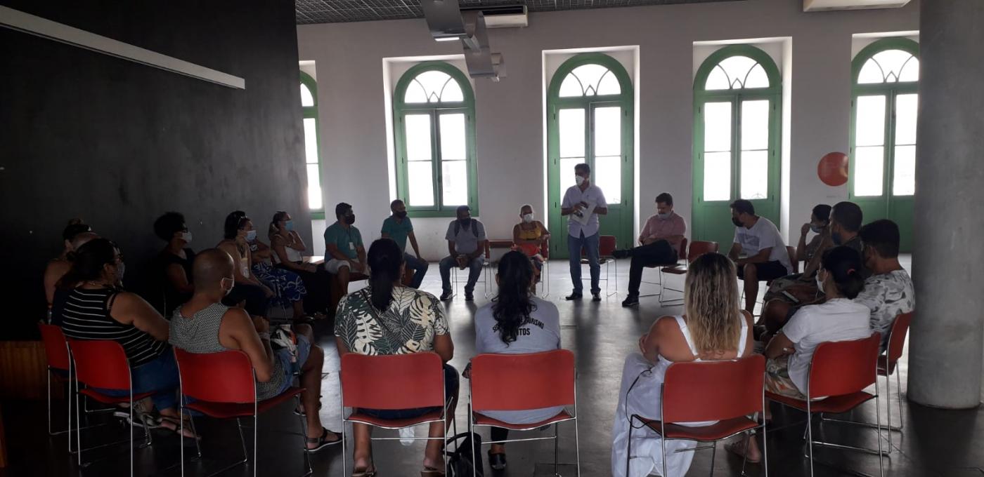 pessoas estão reunidas em roda, sentadas. Ao fundo, um homem em pé fala com todos. O ambiente é uma das salas do Museu Pelé, com grandes portas-balcões. #paratodosverem