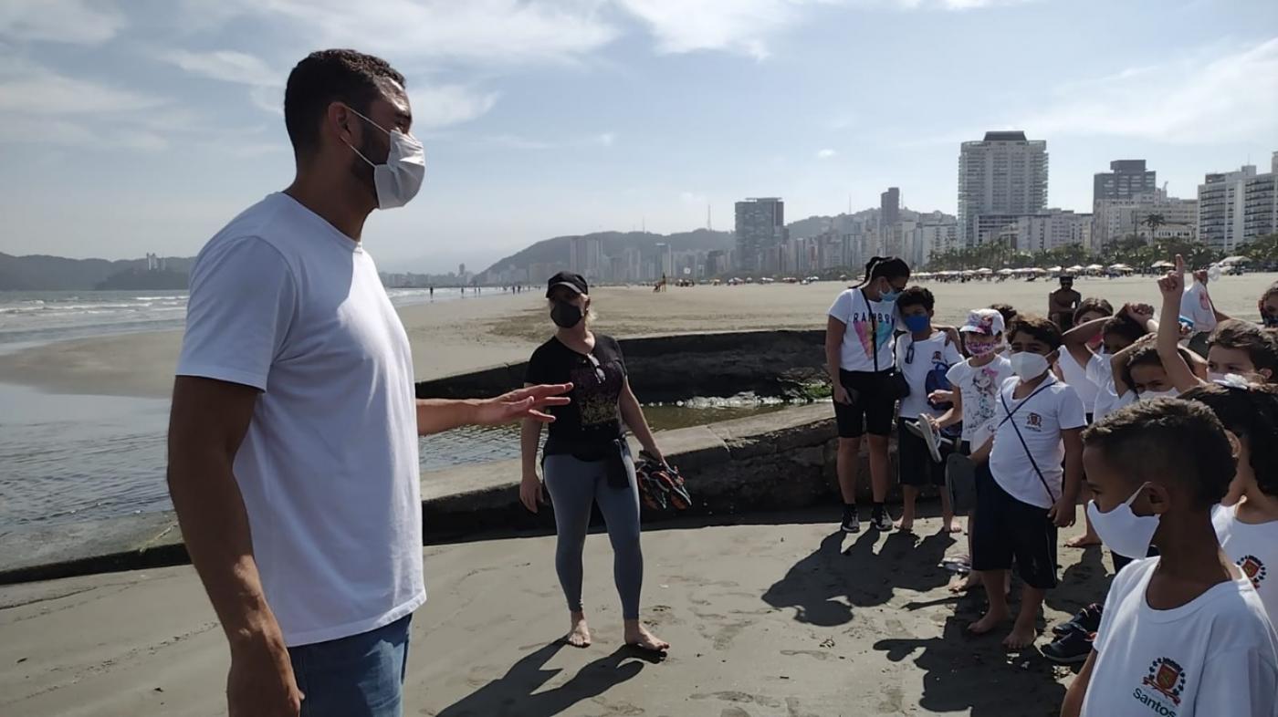 professor conversa com alunos na beira da água na praia e próximo a canal. #paratodosverem