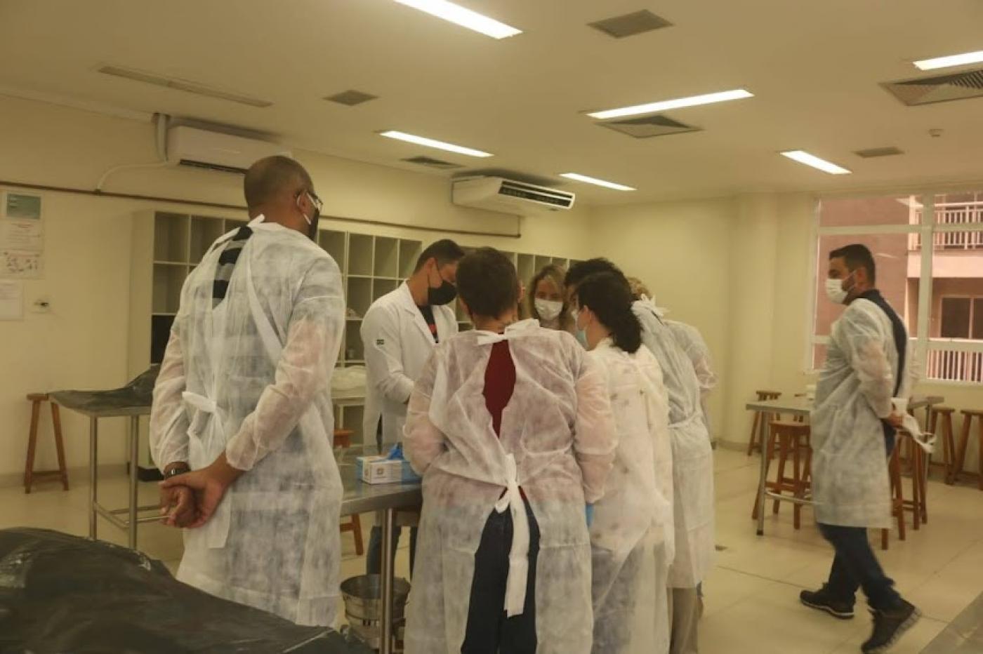 pessoas vestindo avental branco estão em pé em torno de uma mesa em uma sala. Não é possível ver o que há sobre a mesa. #paratodosverem