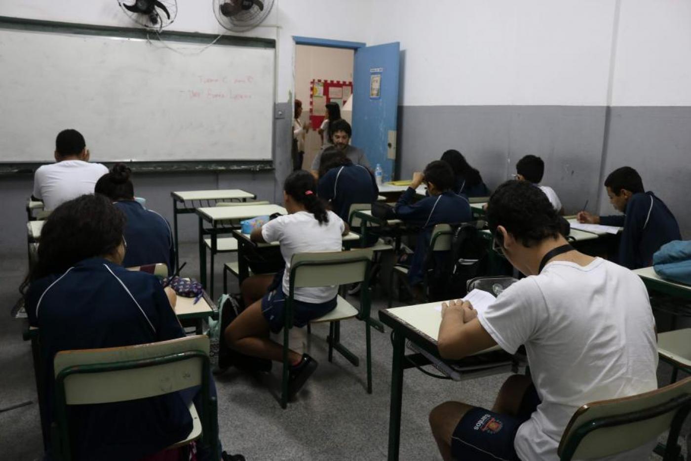 Alunos estão sentados em sala de aula. Todos estão de costas para a foto e de frente para a lousa. #Paratodosverem