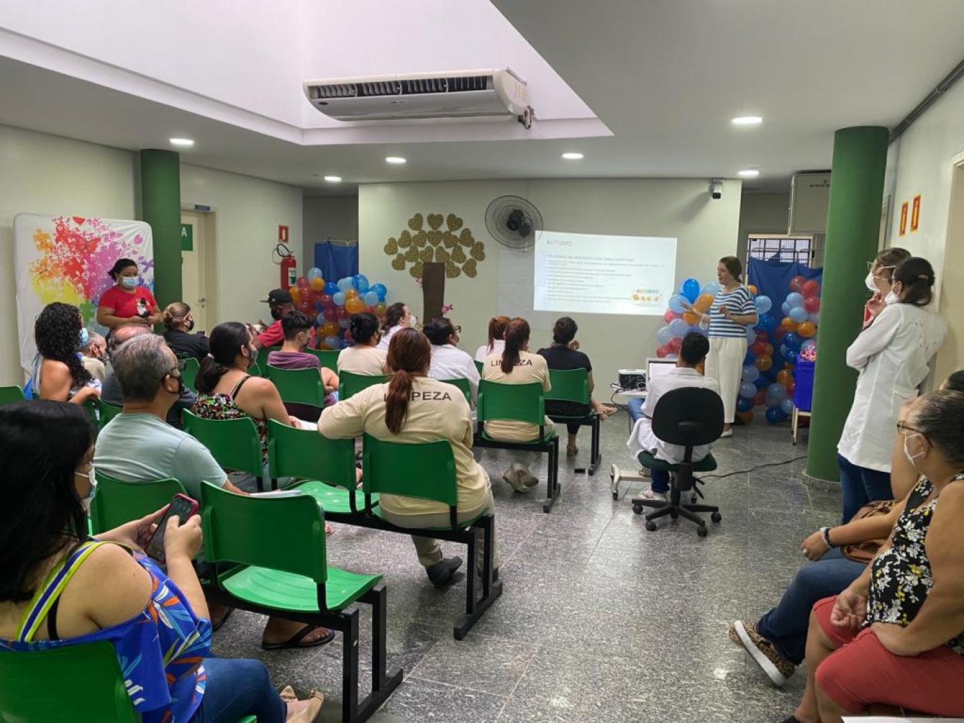 público está sentado em sala assistindo palestra. Ao fundo, uma imagem é projetada na parede. A sala está decorada com balões coloridos. #paratodosverem