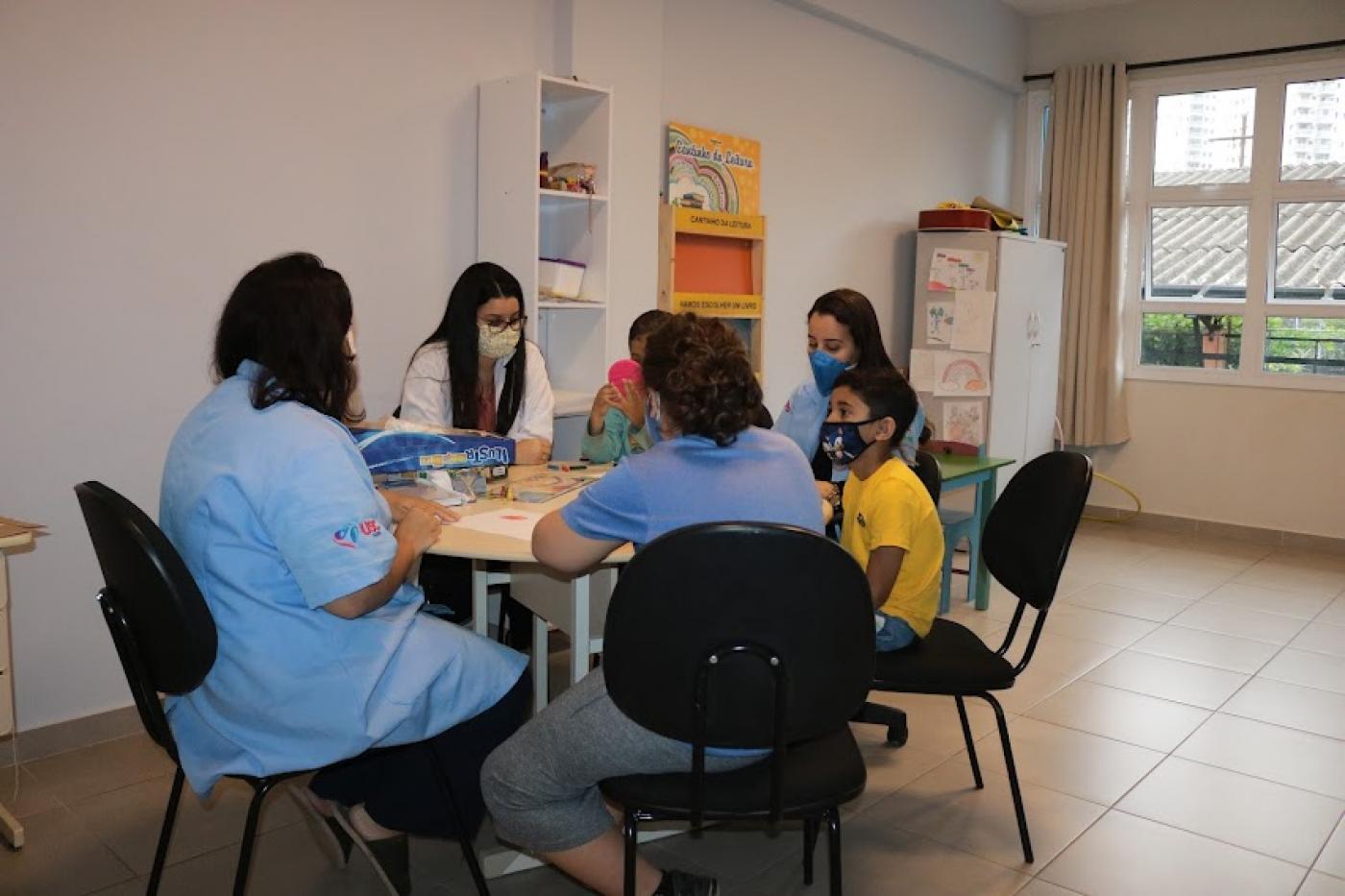 várias pessoas sentadas em torno de uma mesa. Há crianças, jovens e adultos. Ambiente é de uma escola. #paratodosverem