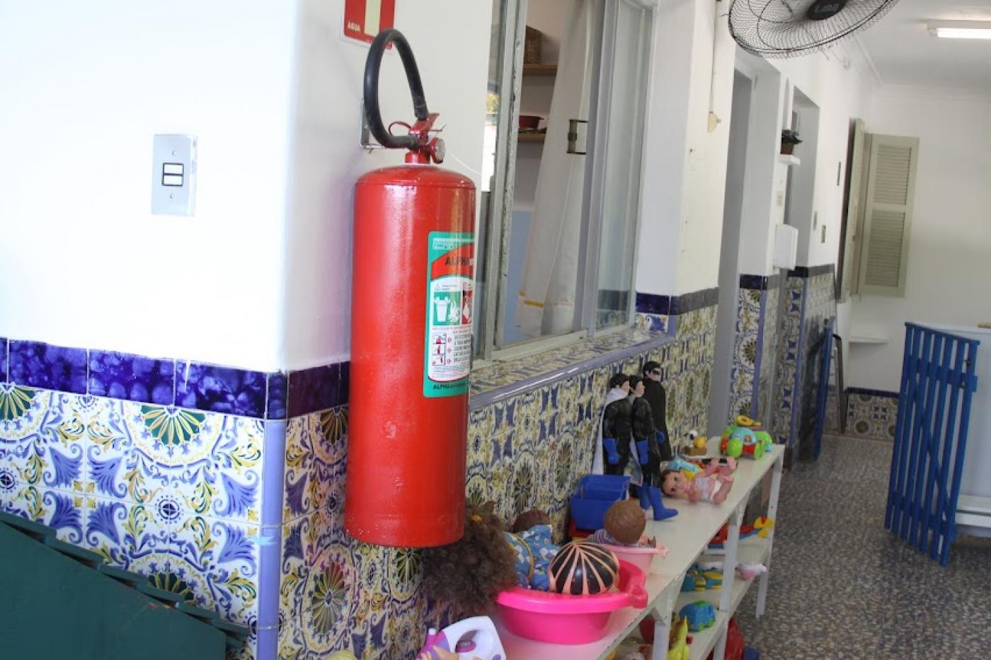 extintor de incêndio afixado em parede de escola. Abaixo, um móvel aberto, com vários recipientes com brinquedos. #paratodosverem