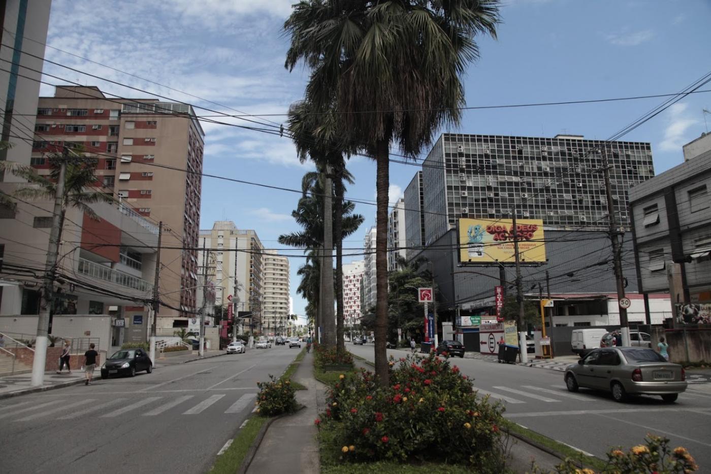 Avenida larga, com canteiro central arborizado com palmeiras, dividindo as duas pistas. #Paratodosverem