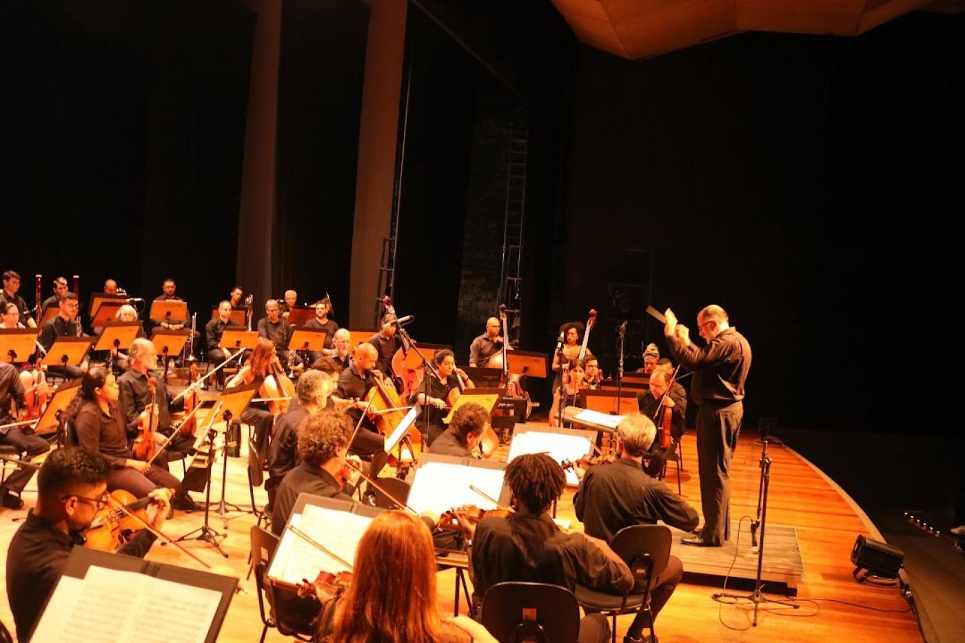 os músicos da orquestra no palco e o regente à frente. #paratodosverem 