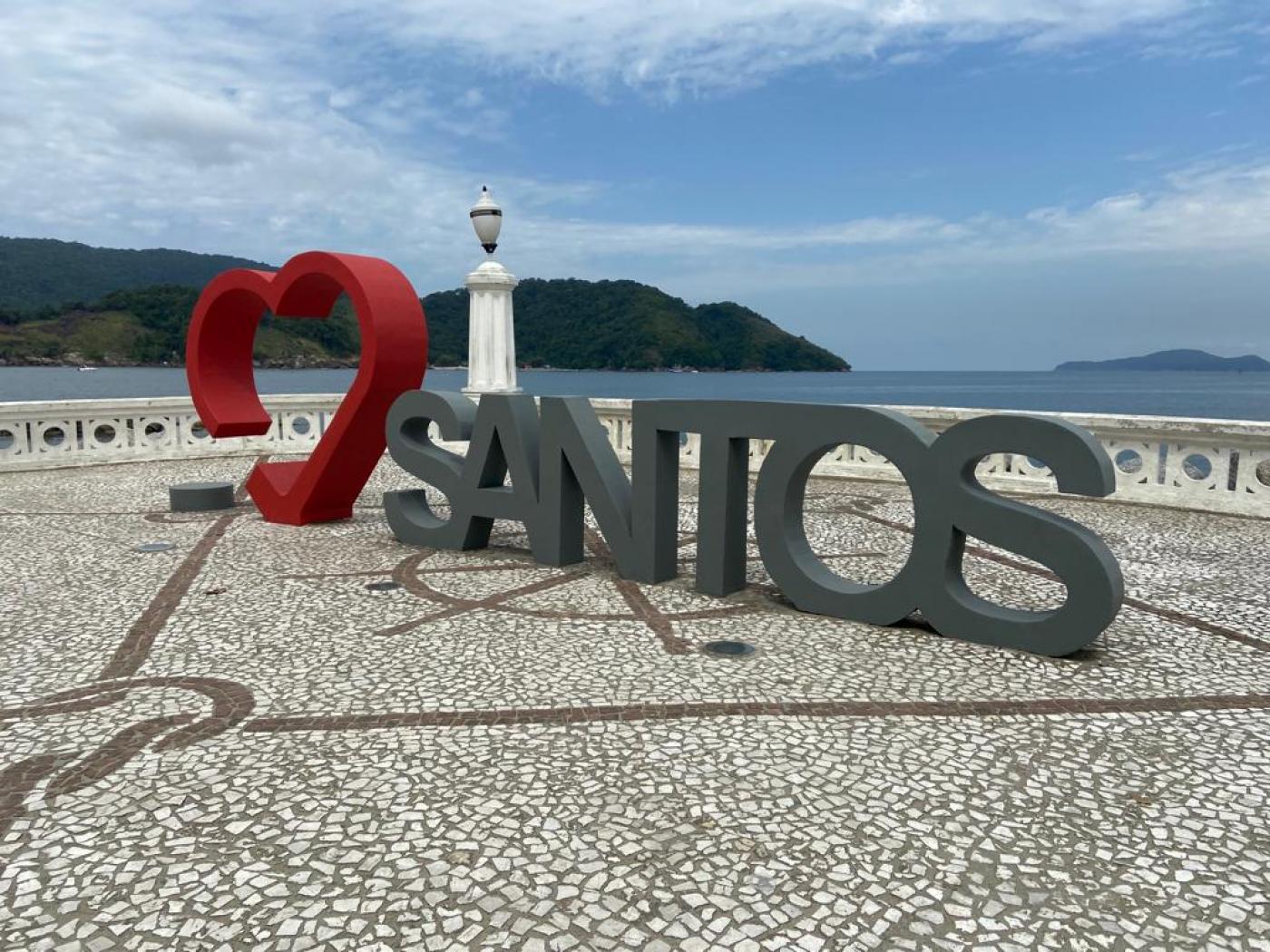 a escultura em um recuo na orla, com o coração em vermelho ao fundo e a palavra santos em cinza. #paratodosverem