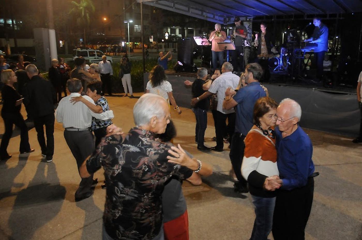 pessoas estão dançando em pista na orla da praia. Banda está tocando ao fundo sob tenda. #paratodosverem