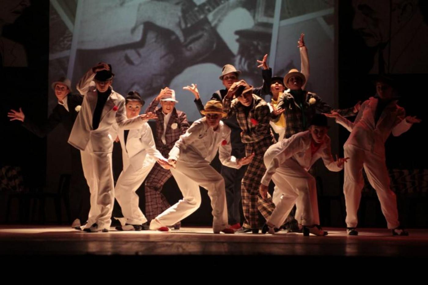 Bailarinos estão no palco fazendo coreografia. Eles usam calças, ternos e chapéus. #Paratodosverem