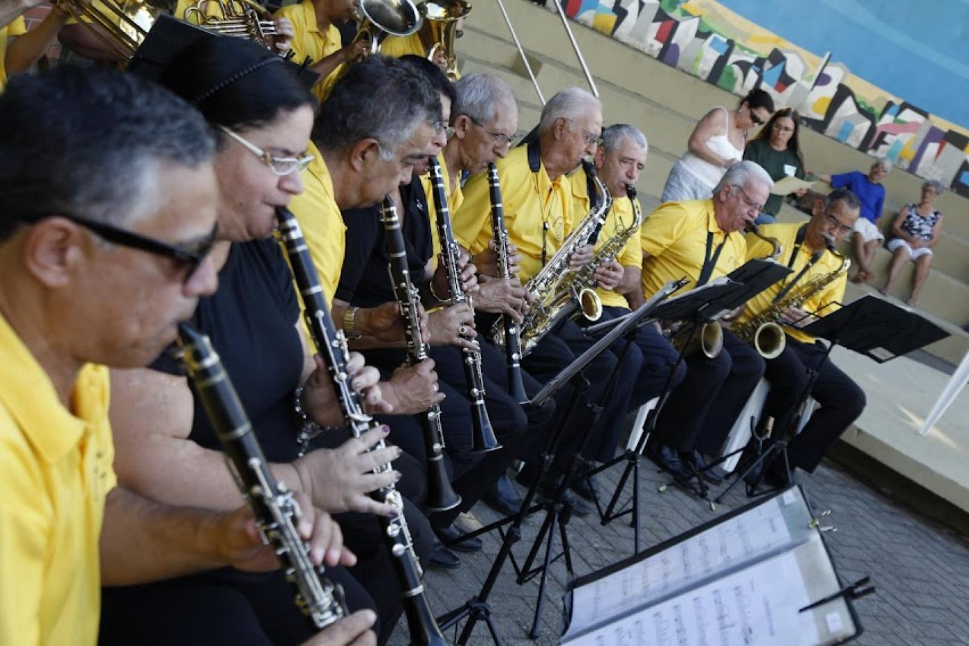 close de músicos sentados lado a lado tocando instrumentos de sopro. eles usam camisetas da mesma cor, em amarelo. Diante deles há estantes com partituras. #paratodosverem