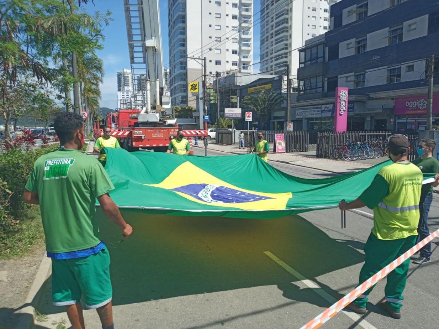 homens estendem a bandeira na rua preparando o hasteamento. #paratodosverem