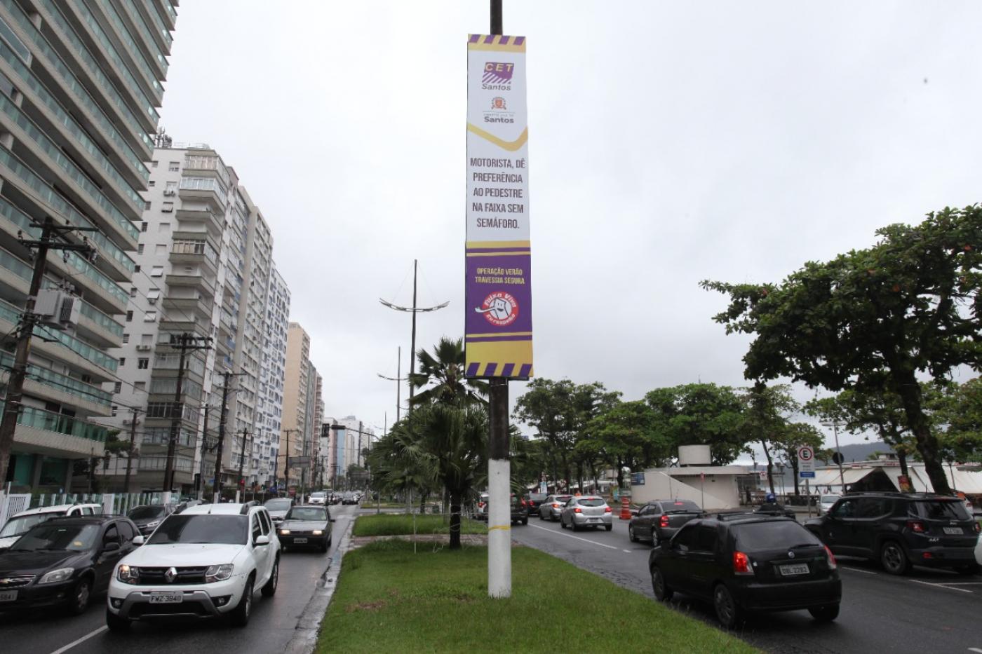 banner afixado em poste em canteiro central de avenida da orla. Carros passam nas duas pistas, indo e vindo. #paratodosverem