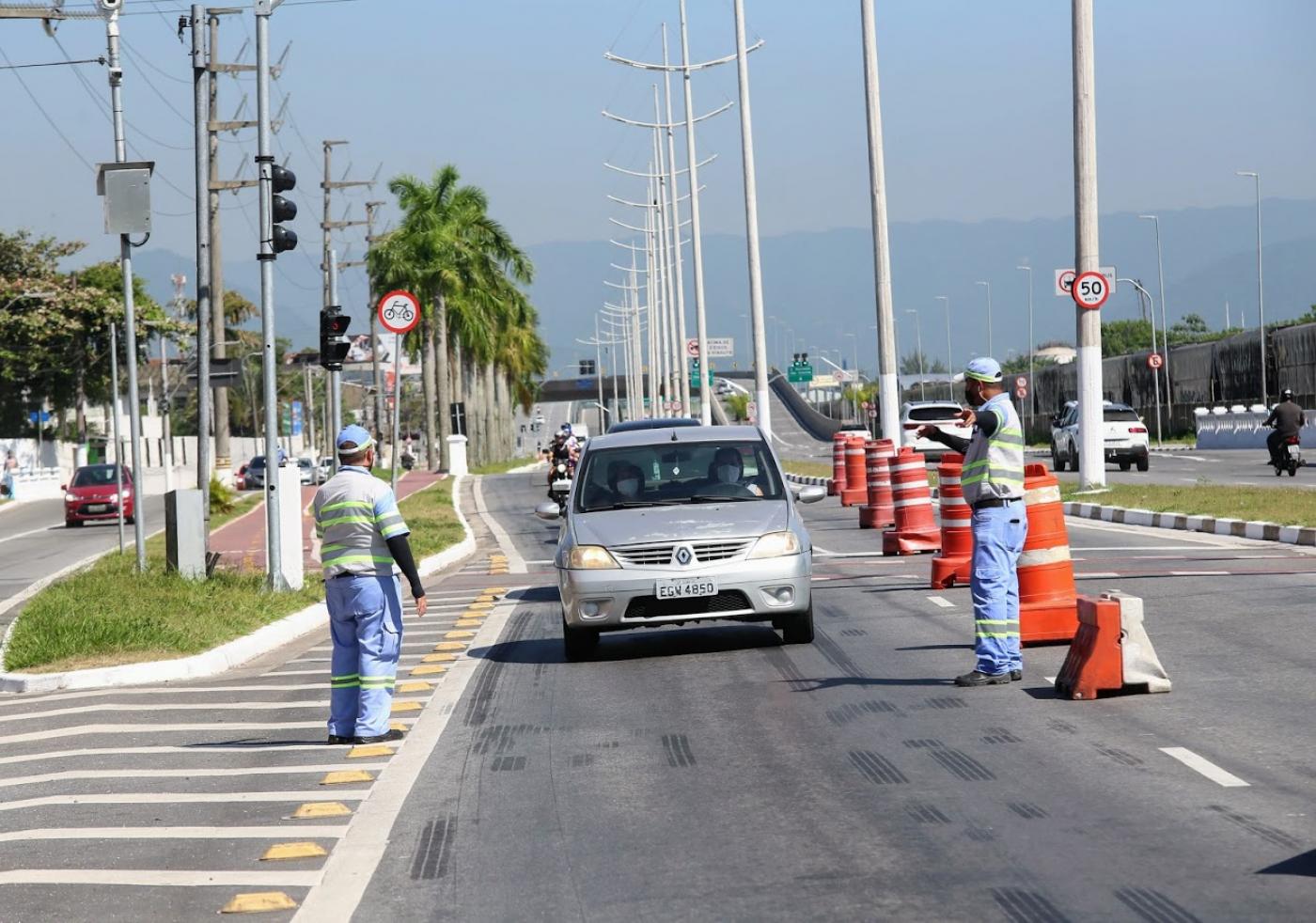 Dois agentes da CET em cada lado de uma vista de veículos. Uma fila de carros está formada e passando ao lado de cones. #paratodosverem