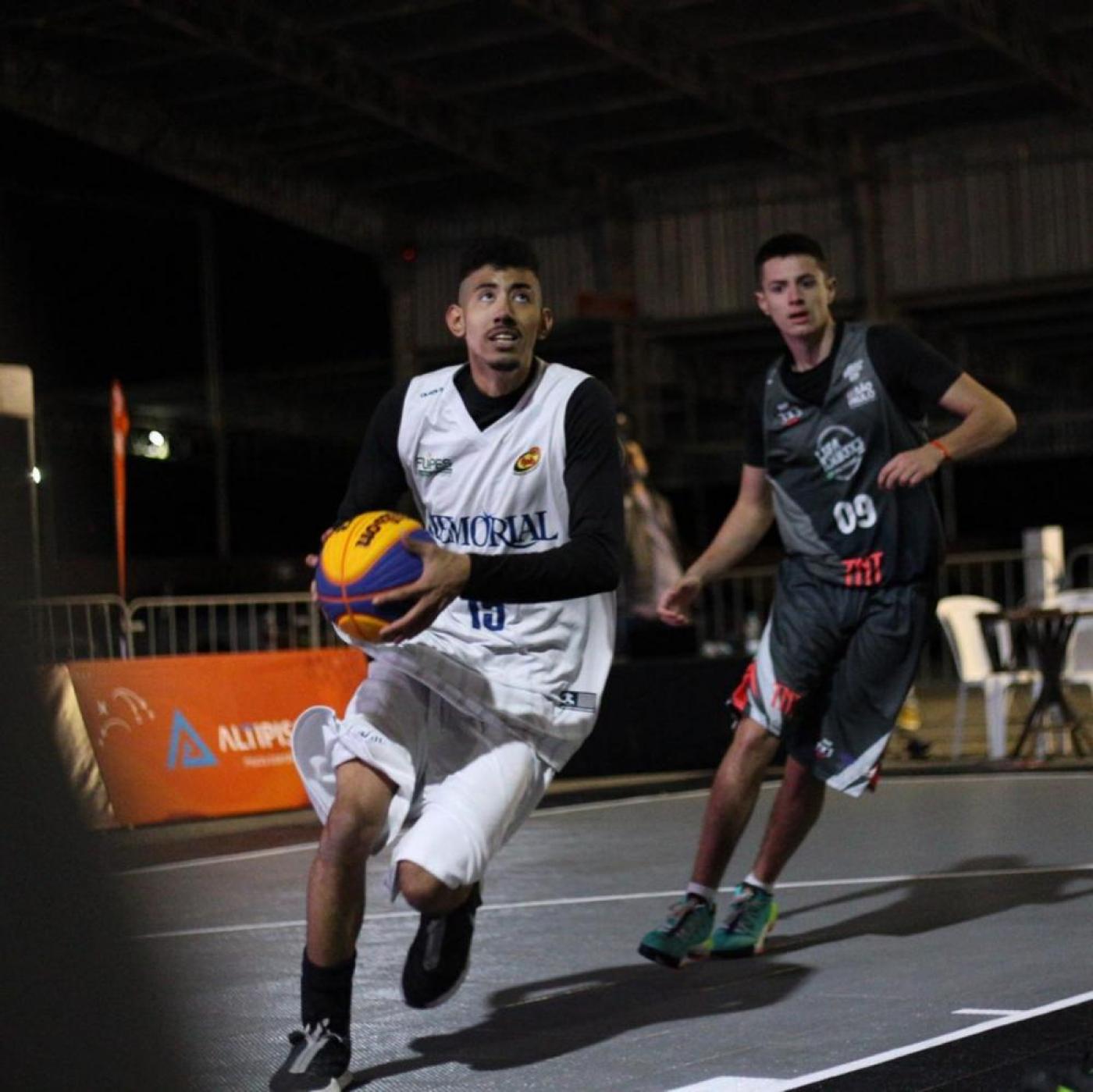 Jogador da equipe de Santos, com uniforme branco, segura a bola de basquete e olha para o alto, com adversário acompanhando de perto. #pracegover