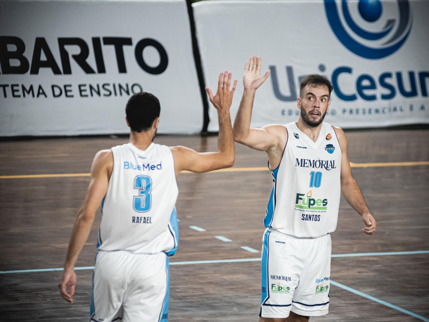 dois jogadores estão em quadra eles vão ao encontro um do outro e batem as mãos no alto. Um está de costas para a foto e o outro, de frente. #paratodosverem