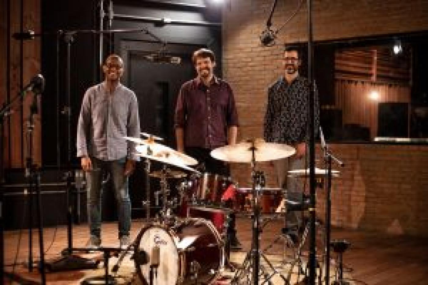 Integrantes do Mário Gaiotto Trio posam para foto atrás de bateria montada em estúdio de música. #paratodosverem