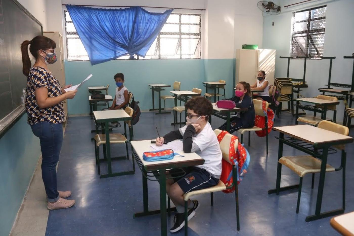 Em uma sala de aula, professora lê um papel e alunos a observam sentados nas suas carteiras - #paratodosverem