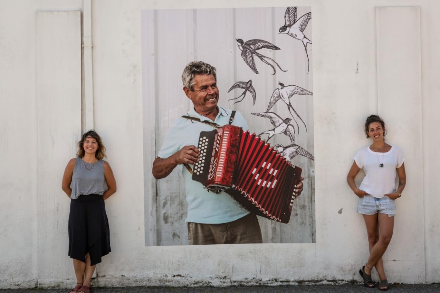 Beatriz e Carolina estão em pé ao lado da imagem de um homem tocando concertina. #Paratodosverem