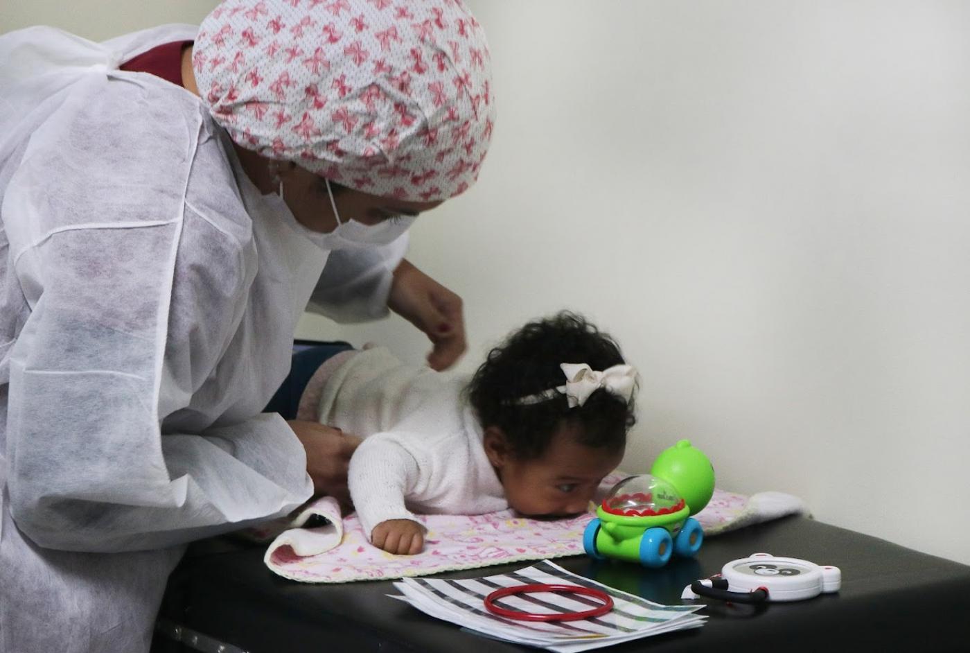 mulher segura bebê que está deitada de bruços observando um brinquedo colorido. #paratodosverem