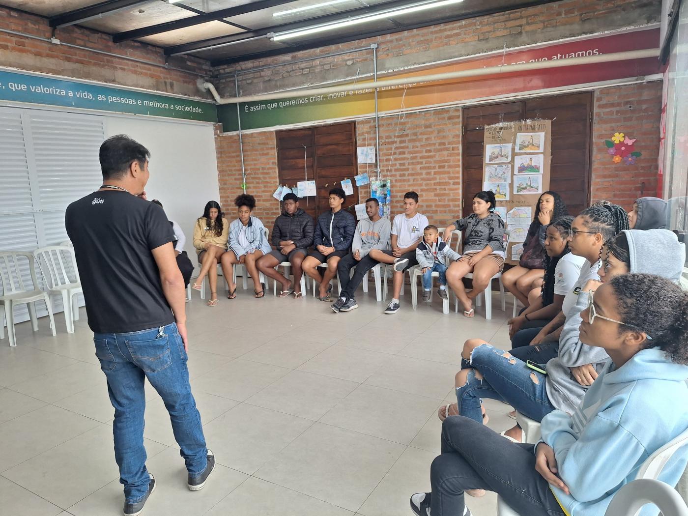 homem está em pé, de costas para a foto, falando a grupo de jovens que estão sentados em um auditório. #paratodosverem 