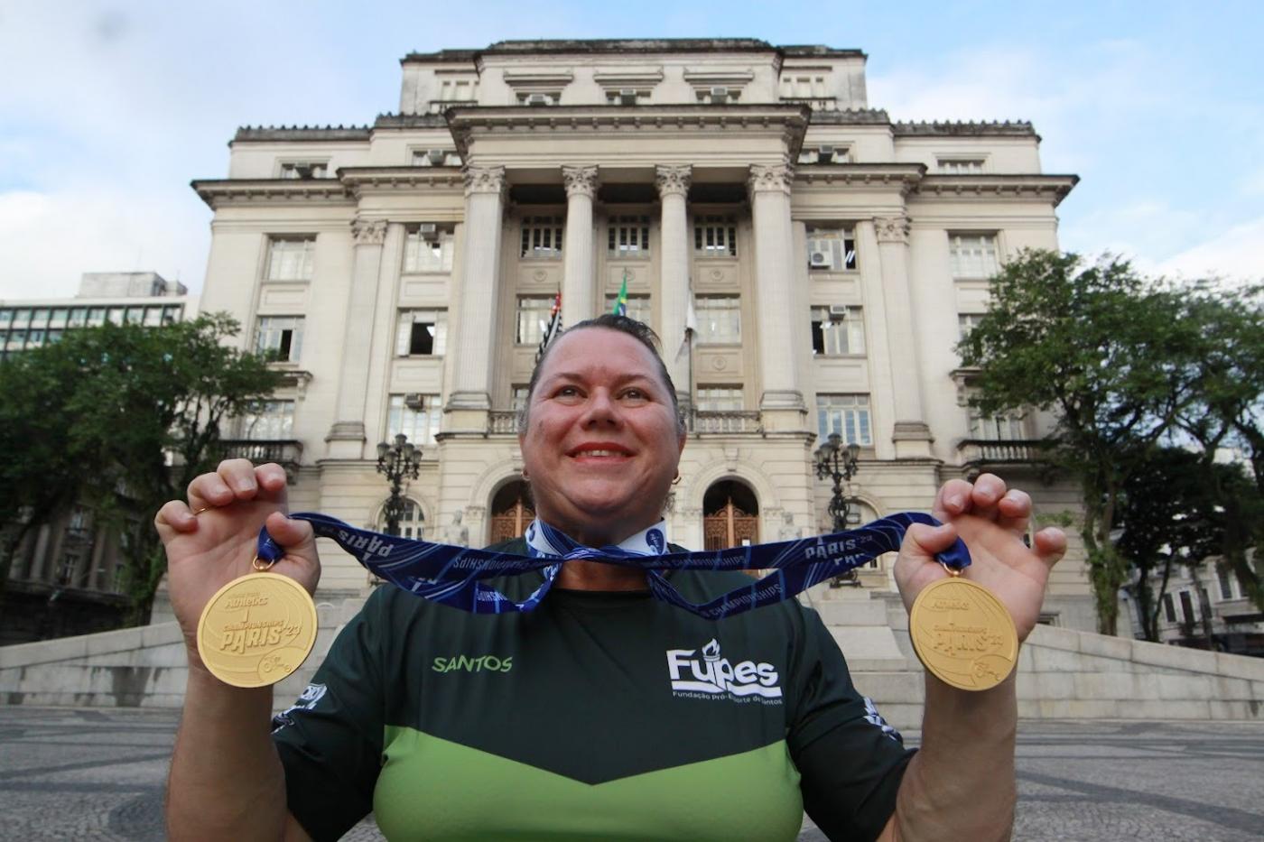 beth gomes mostra duas medalhas na frente da prefeitura #paratodosverem