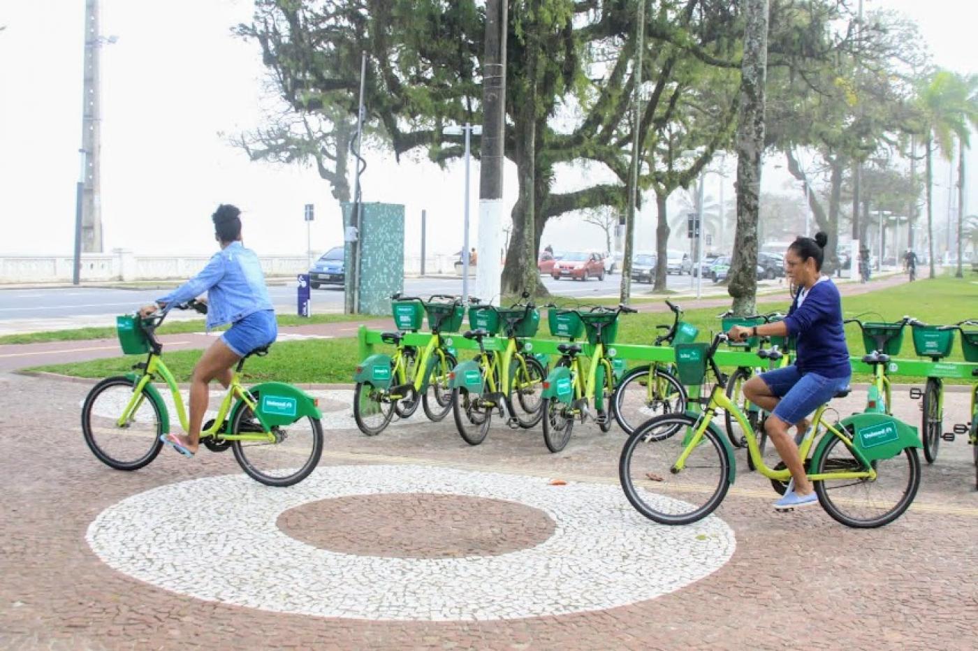 duas mulheres estão montadas em bicicletas do bike santos ao lado de estação.  A estação fica em uma faixa de jardim da orla. #paratodosverem