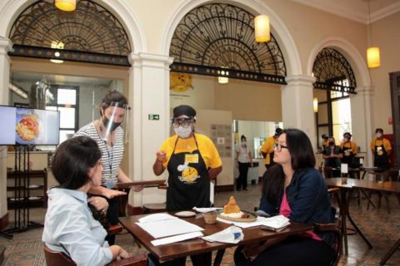 Duas mulheres, sentadas, são atendidas no Restaurante-Escola. #pracegover