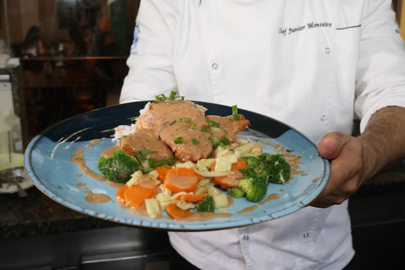 mãos do chef junior monteiro seguram prato com peixe frito, legumes e arroz. #paratodosverem