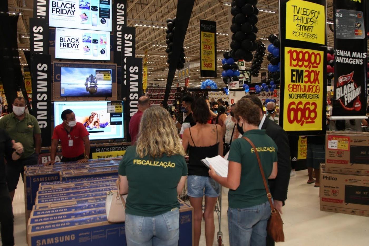 Fiscais identificadas por camisetas estão de costas para a foto em loja de eletrodomésticos. O ambiente está cheio de caixas e cartazes com promoções expostos. #Paratodosverem