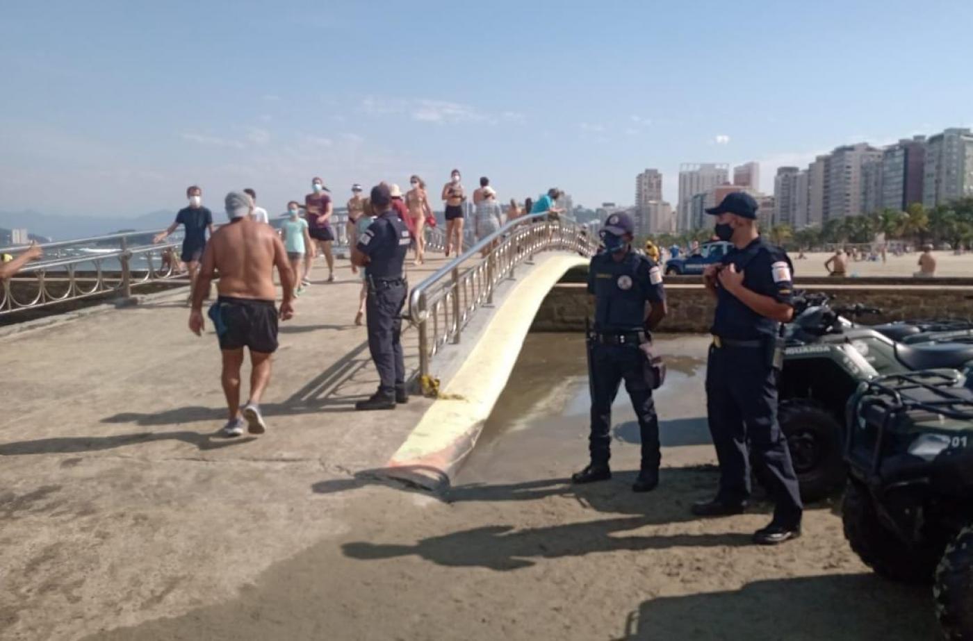 três guardas municipais e quadriciclos estão ao lado de ponte que atravessa canal na faixa de areia. Pessoas cruzam a ponte. #paratodosverem