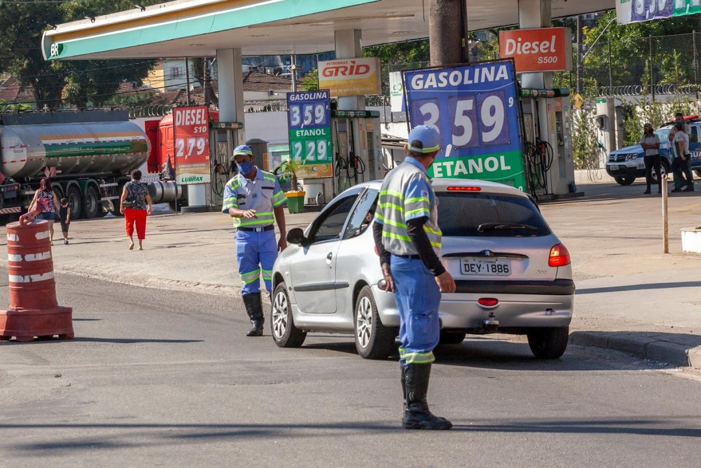 Veículo para próximo à guia de calçada diante de agente da CET. Outro agente está em primeiro plano olhando para o carro. Ao lado direito há um posto de combustível.. #Paratodosverem