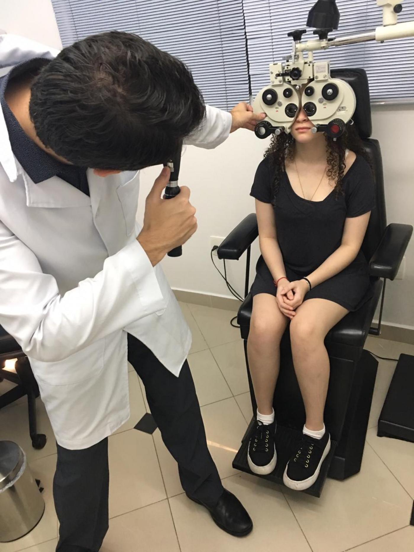Oftalmologista em pé examina aluna sentada com aparelhos nos olhos. #pracegover