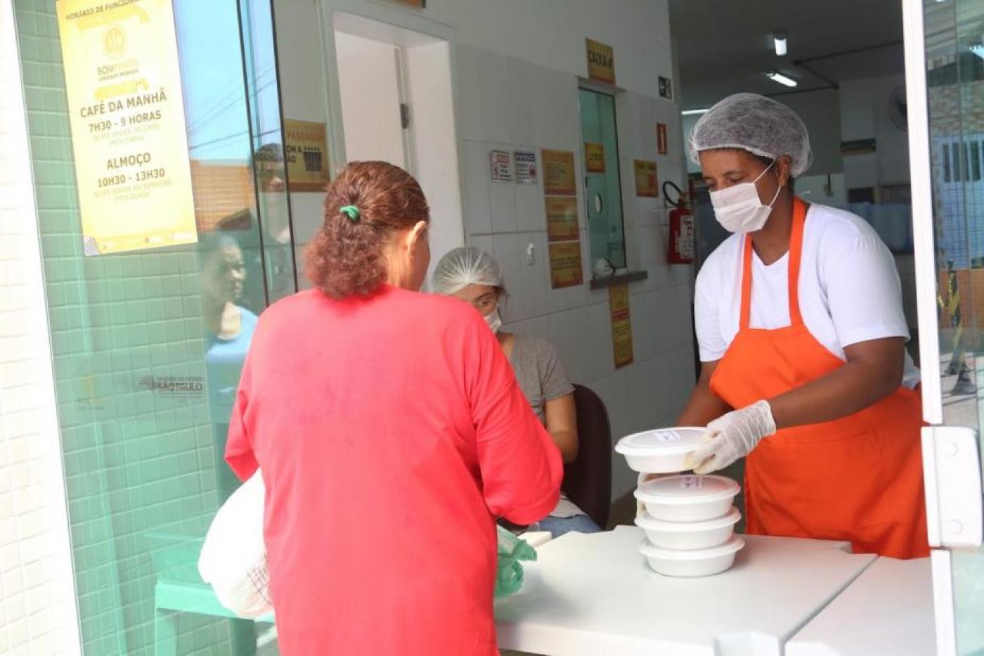 Mulher entrega marmita para outra mulher de costas. #paratodosverem