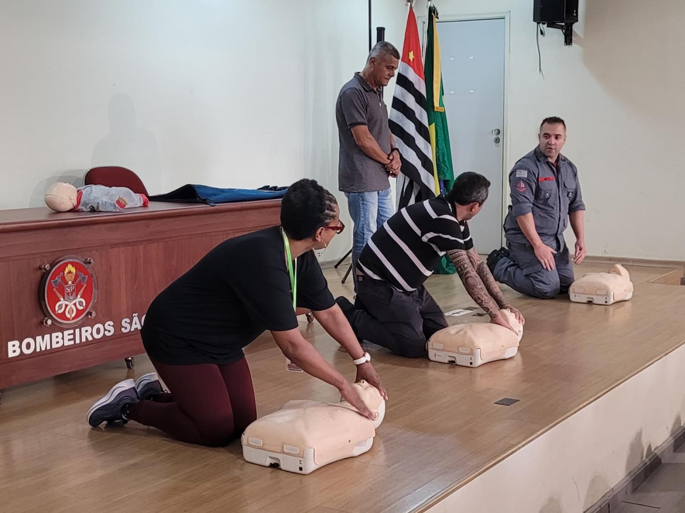 três pessoas estão ajoelhadas no chão massageando peito de boneco que simula uma pessoa. Uma dessas pessoas é o bombeiro que orienta o procedimento. #paratodosverem 