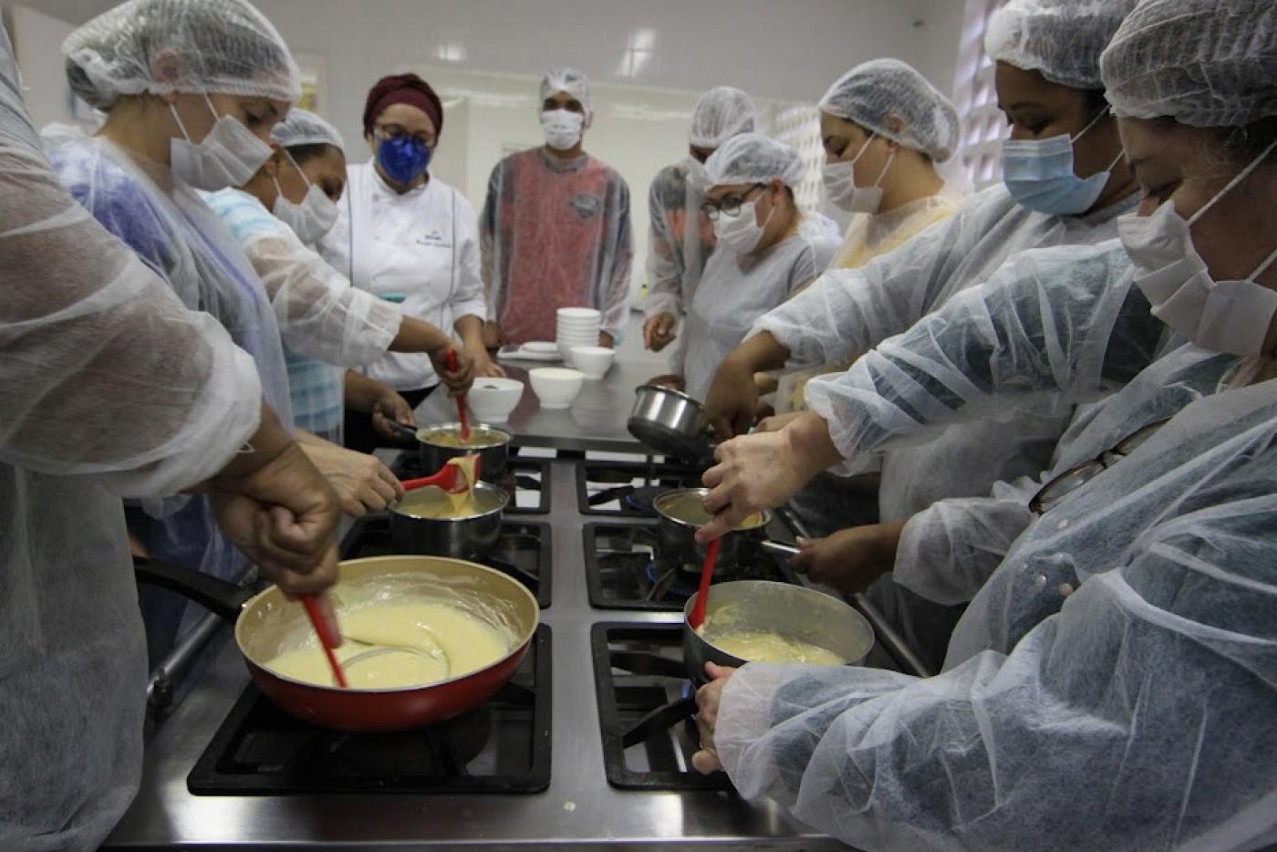 várias pessoas estão mexendo em panelas, fazendo receita de brigadeiro, em torno de um fogão industrial. #paratodosverem