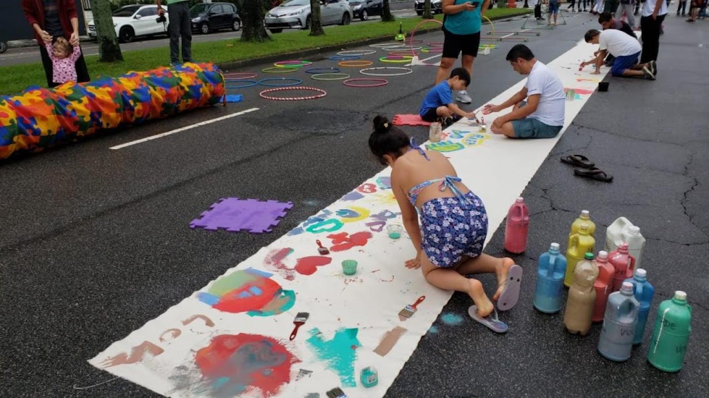 avenida aberta somente para brincadeiras, com uma grande faixa de lona sobre a pista. Crianças e adultos pintam a lona com tinta colorida. Ao fundo há bambolês no chão. #paratodosverem