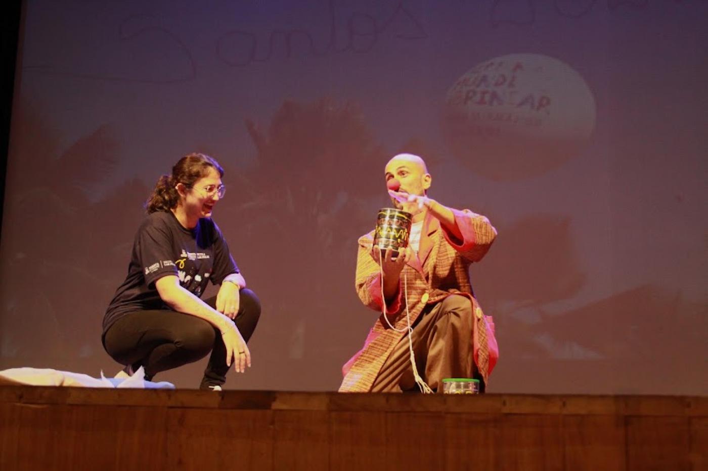 Mulher e palhaço estão lado a lado em palco. eles estão agachados. A mulher ri. #paratodosverem