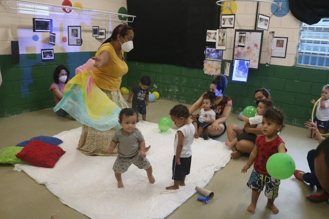 Crianças brincando dentro de uma sala com adulto acompanhando #paratodosverem