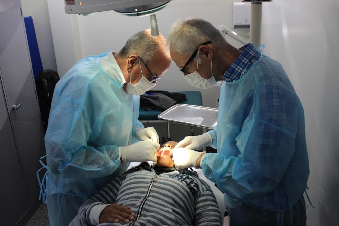 dois cirurgiões estão em pé atendendo um homem que está deitado de boca aberta. #paratodosverem
