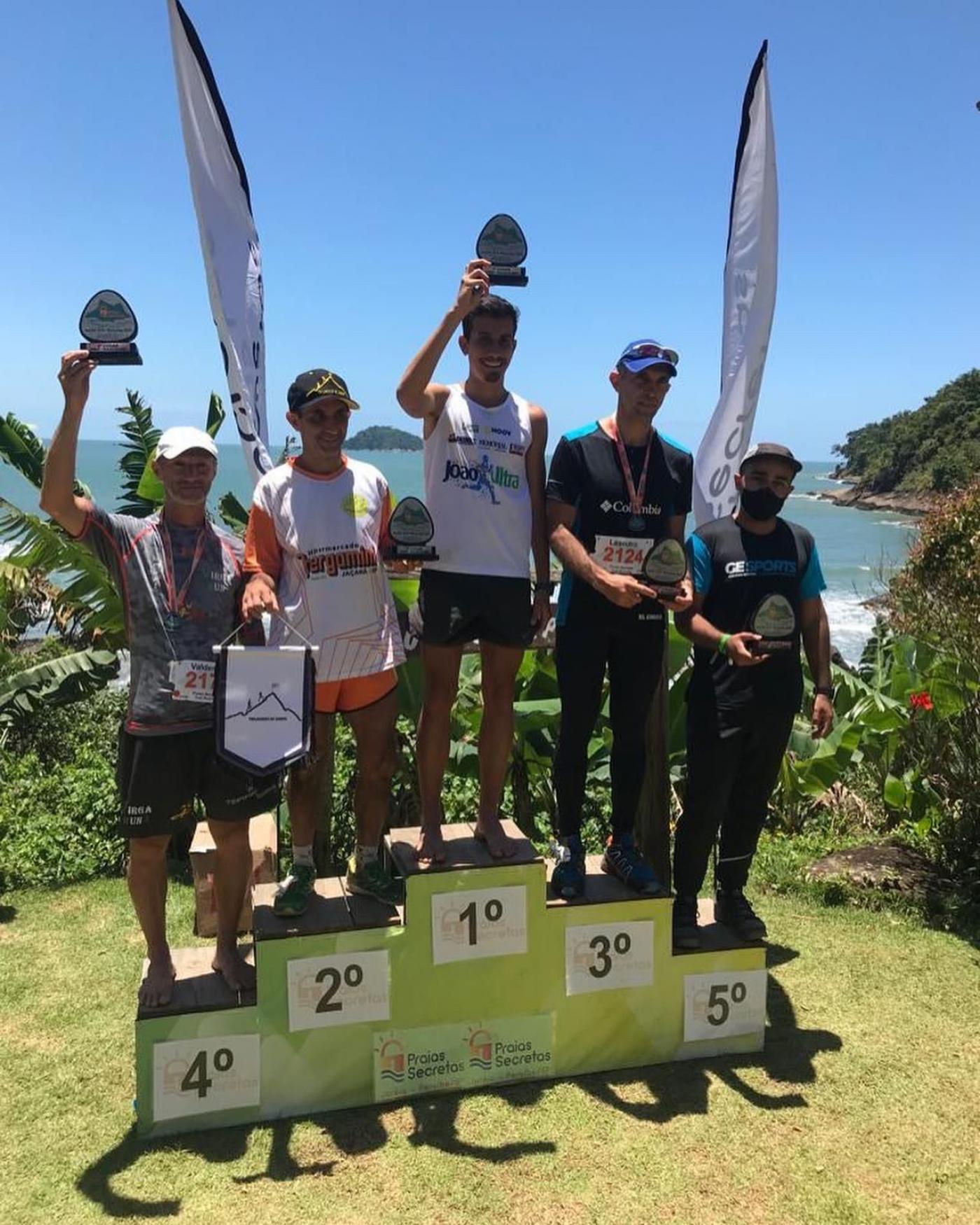 Atletas no pódio erguem troféu com paisagem de fundo #paratodosverem