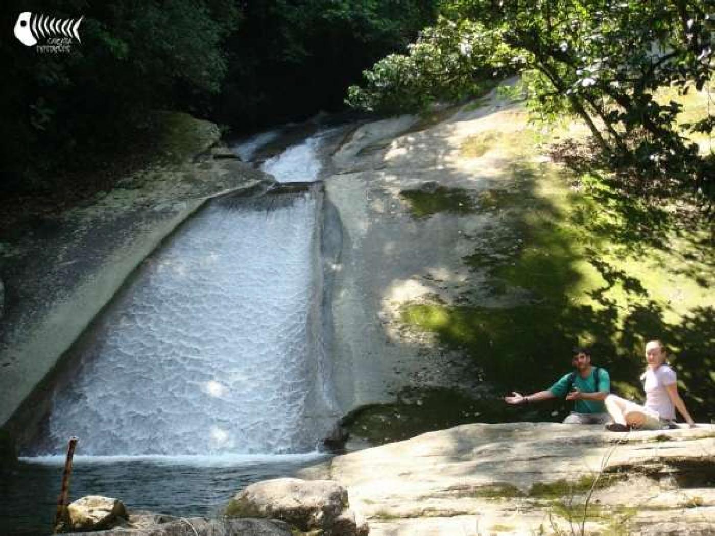 cachoeira com piscina natural na parte inferior. Do lado direito, duas pessoas estão sentadas sobre pedras. Há vegetação em torno. #paratodosverem