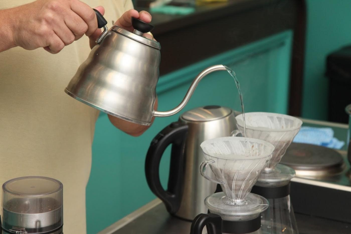 mãos seguram bule com água quente que é despejada em filtro de café que está em coador transparente. Ao lado há outro coador. #paratodosverem 