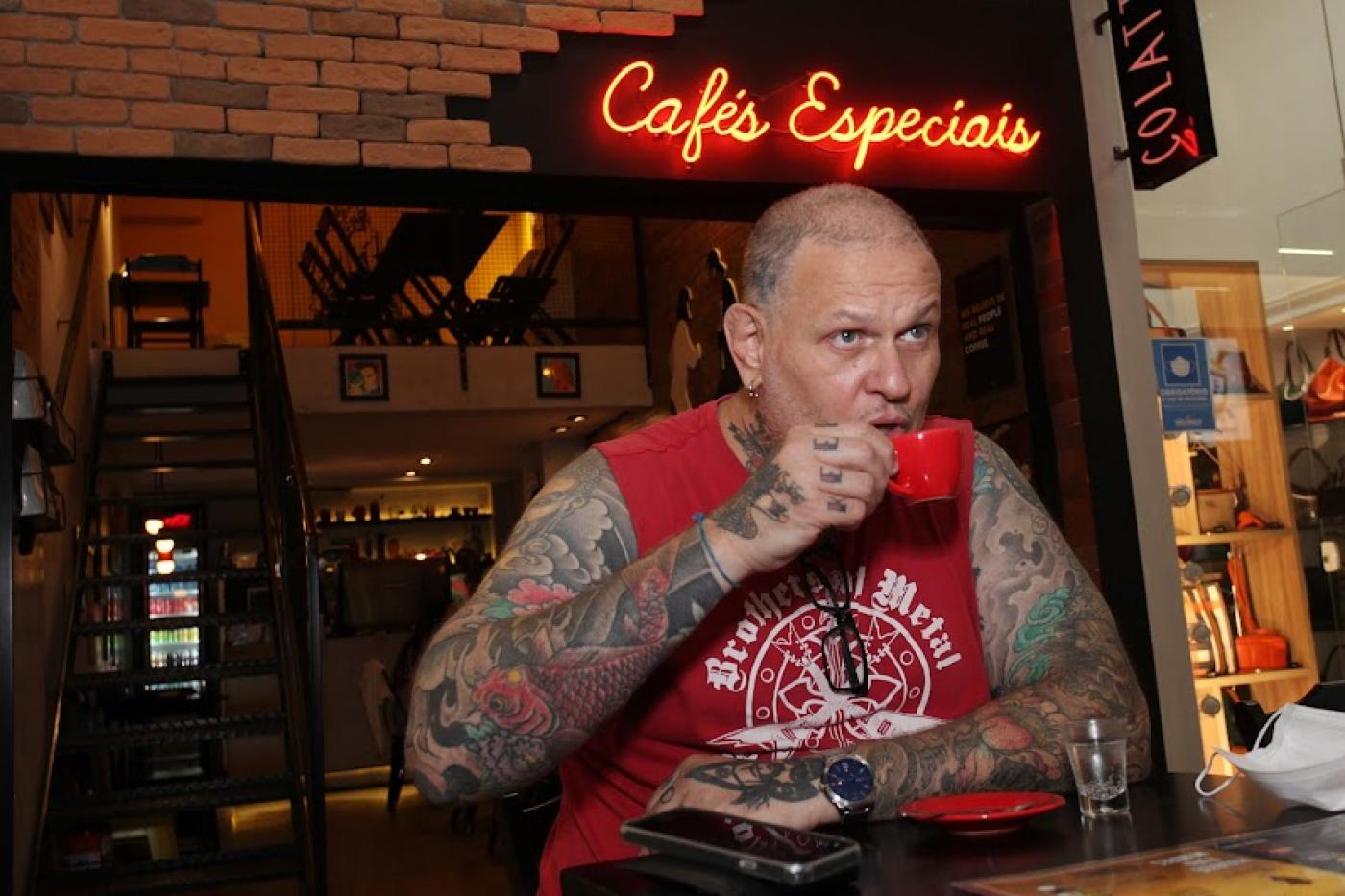 homem coberto de tatuagens nos braços toma café em xícara. Ele está sentado a uma mesa diante de uma cafeteria em cuja fachada tem um luminoso onde se lê Cafés Especiais - #paratodosverem